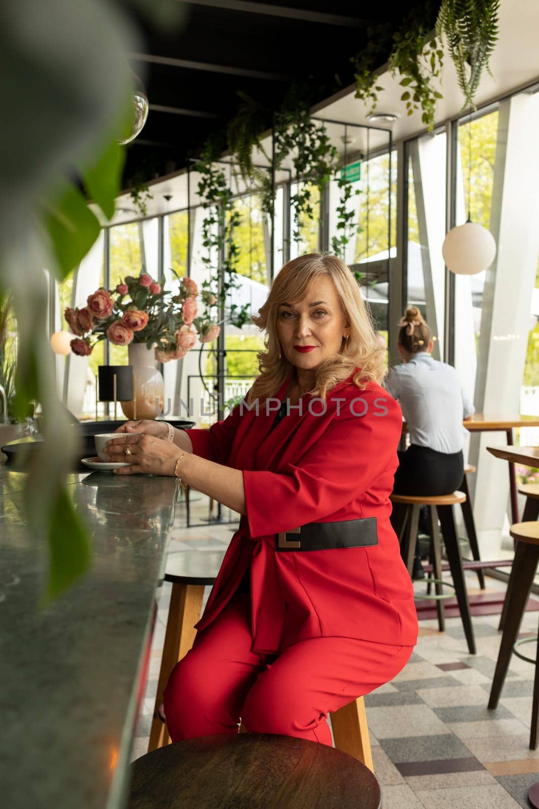 Beautiful Blonde Mature Sexagenarian Woman Drinks Coffee, Tea at Cafe or Restaurant. Modern Senior Female in her 60s wears Red Fashionable Suit, Enjoys Her Life, Time. Happy Retirement. Vertical Plane