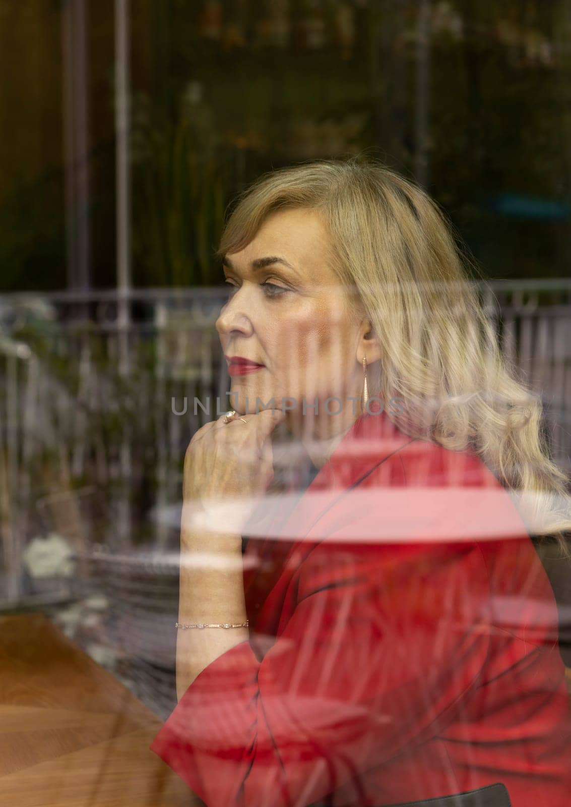 Portrait Beautiful Blonde Mature Sexagenarian Woman Sitting Behind Window Glass of Restaurant or Cafe. Stylish Modern Senior Female in her 60s Thinking About Something. Retirement. Vertical Plane.