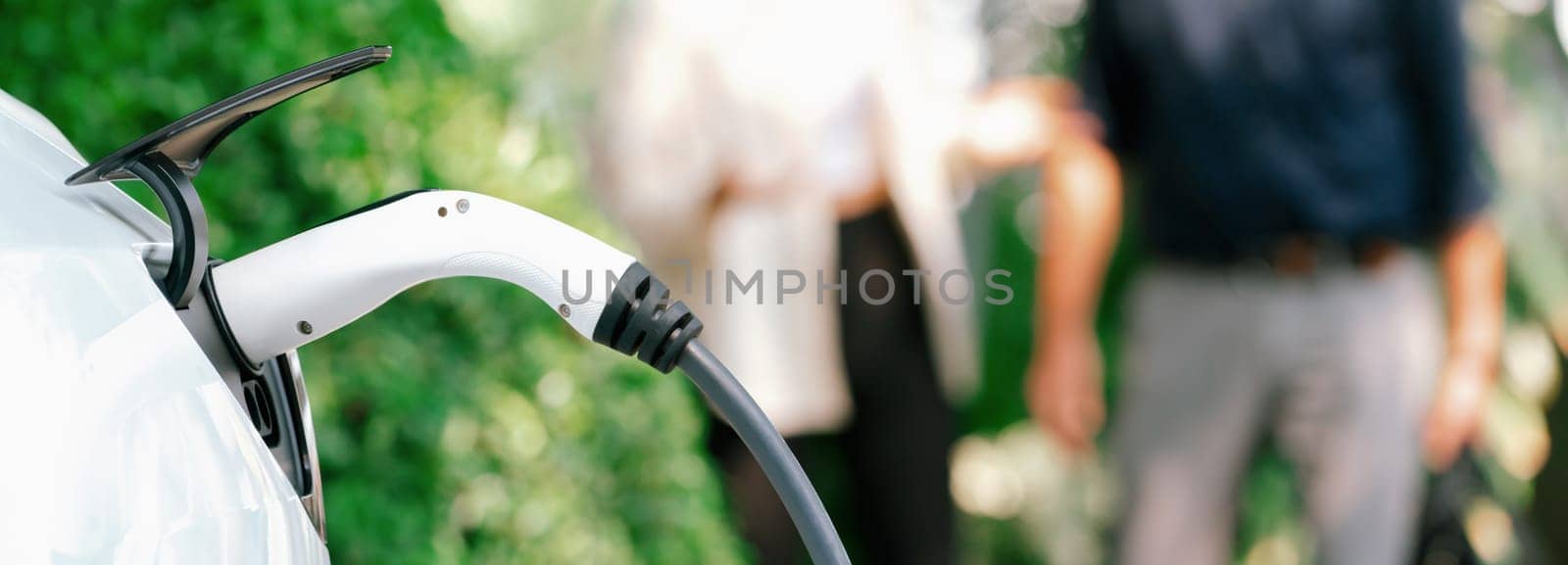 Young couple recharge electric car battery from charging station in green city park in springtime. Rechargeable EV car for sustainable environmental friendly urban travel lifestyle. Panorama Expedient