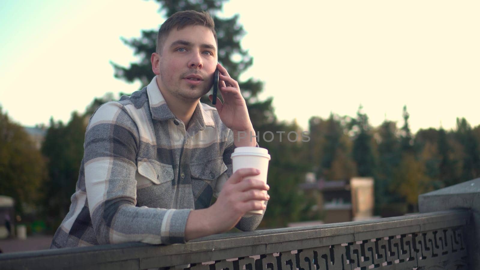 A young man speaks on the phone with a glass of coffee in his hand. A man with a phone near the fence on the embankment with a hot drink in a glass. 4k