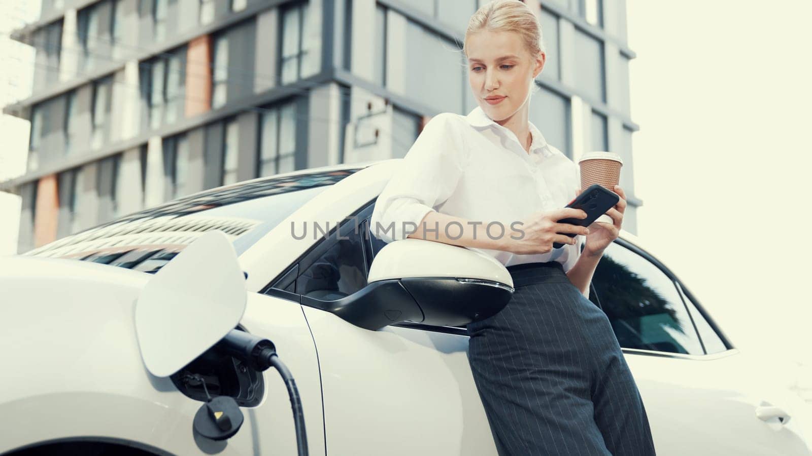 Progressive businesswoman leaning on electric car and charging station before driving around city center. Eco friendly rechargeable EV car powered by sustainable and clean energy.