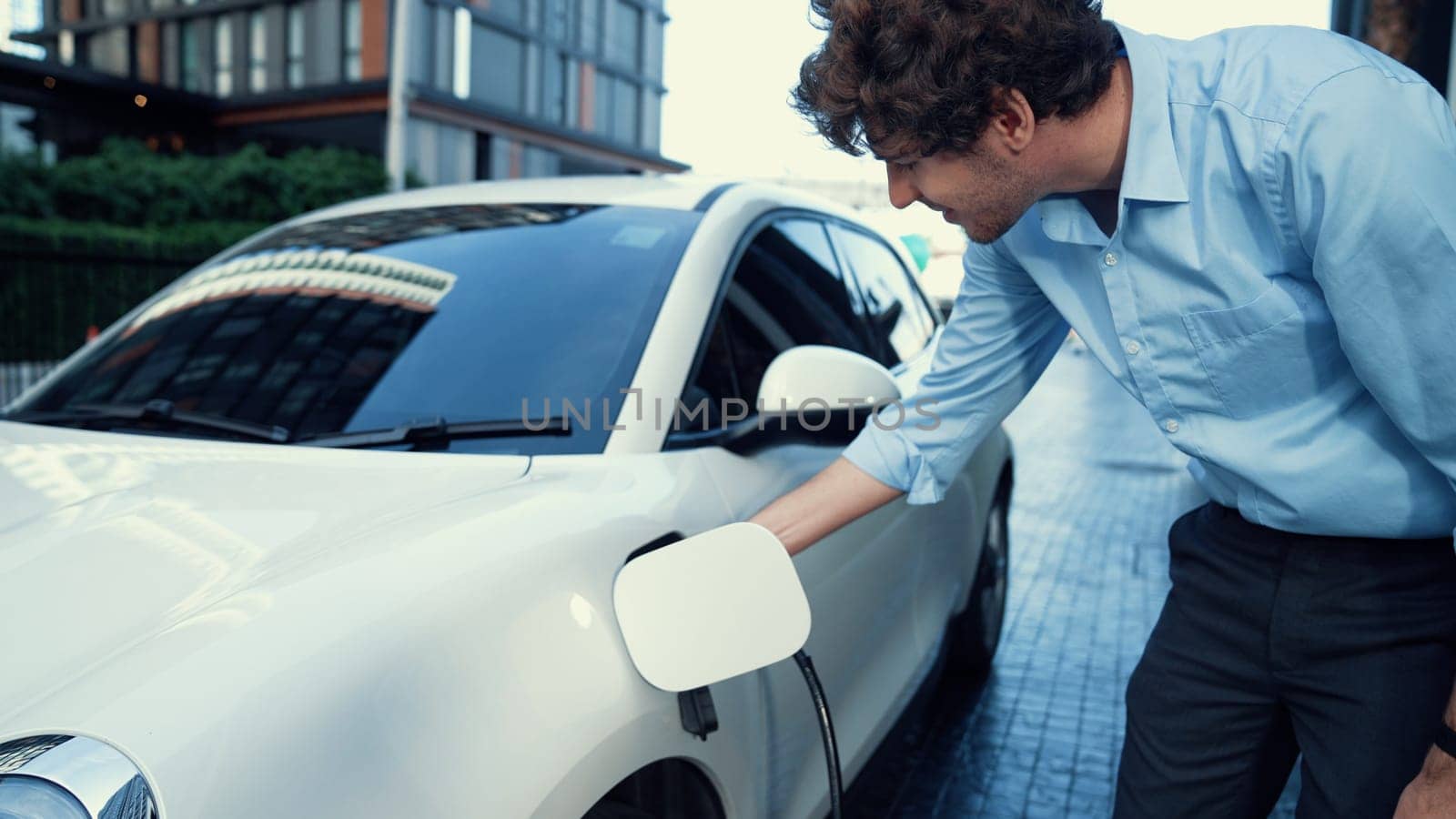 Progressive businessman install charger plug from charging station to his electric car before driving around city center. Eco friendly rechargeable car powered by sustainable and clean energy.