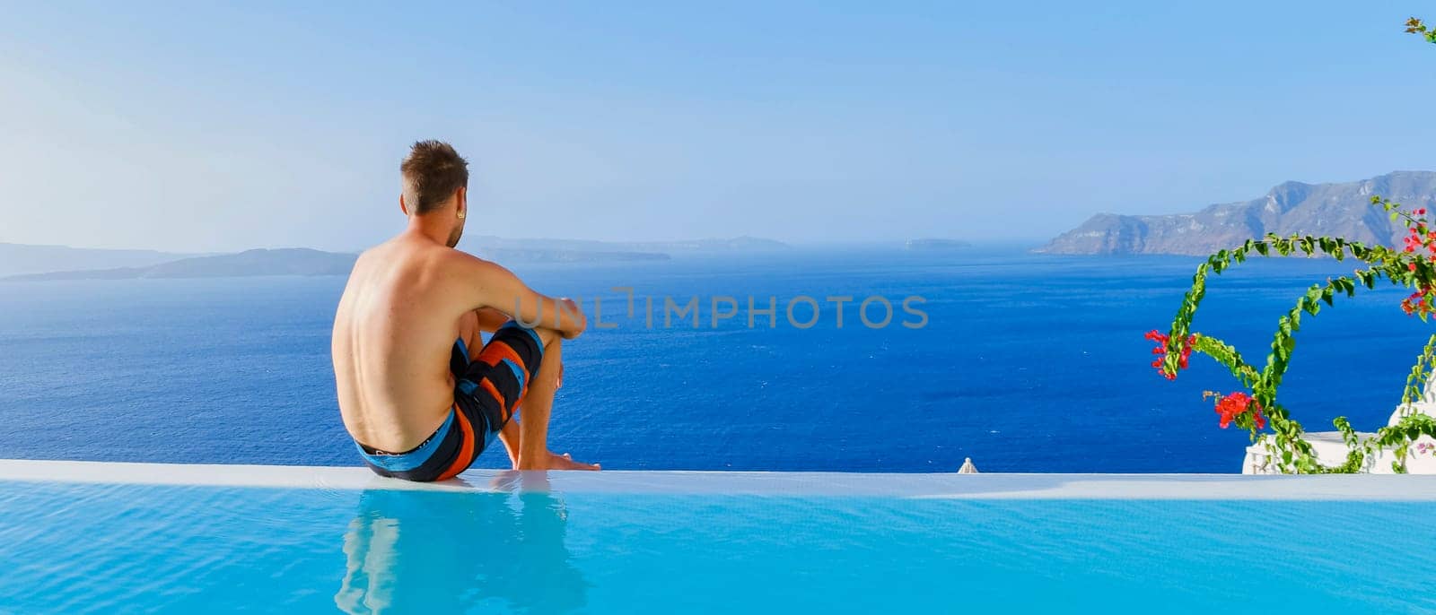 A young man in swim shorts relaxing in an infinity swimming pool during vacation at Santorini, swimming pool looking out over the Caldera ocean of Santorini, Oia Greece, Greek Island Aegean Cyclades