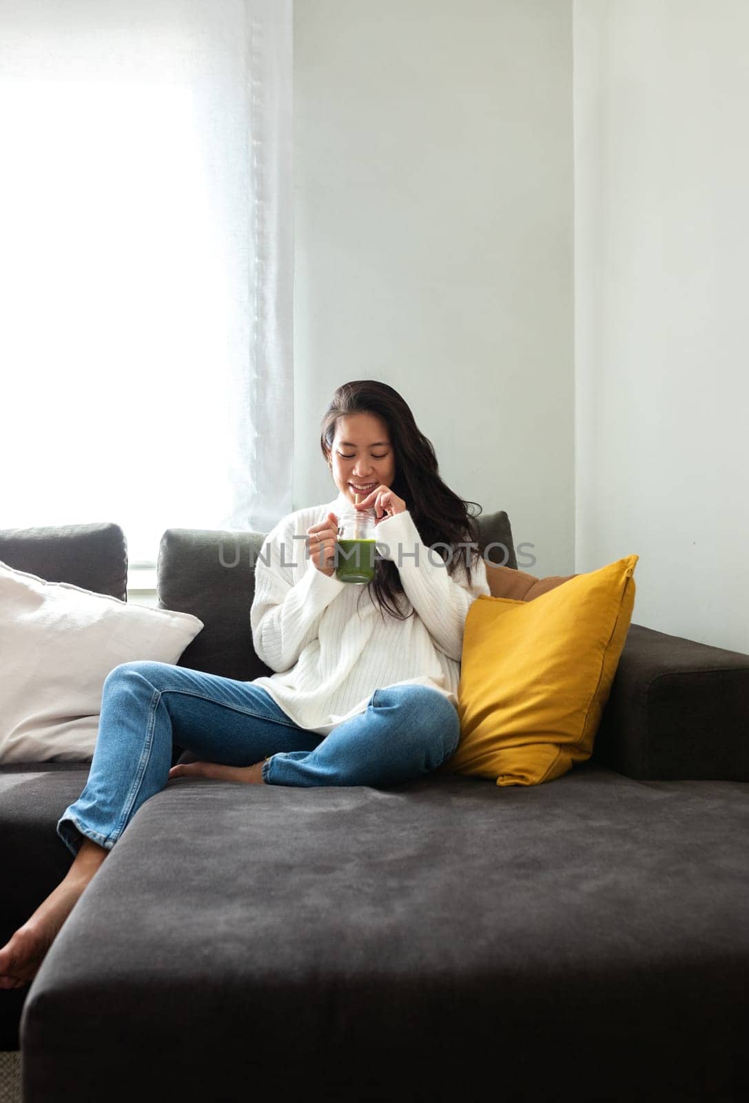 Healthy lifestyle. Young Chinese woman drinking green vegetable juice at home sitting on the sofa. Vertical image. Healthy eating and lifestyle concepts.