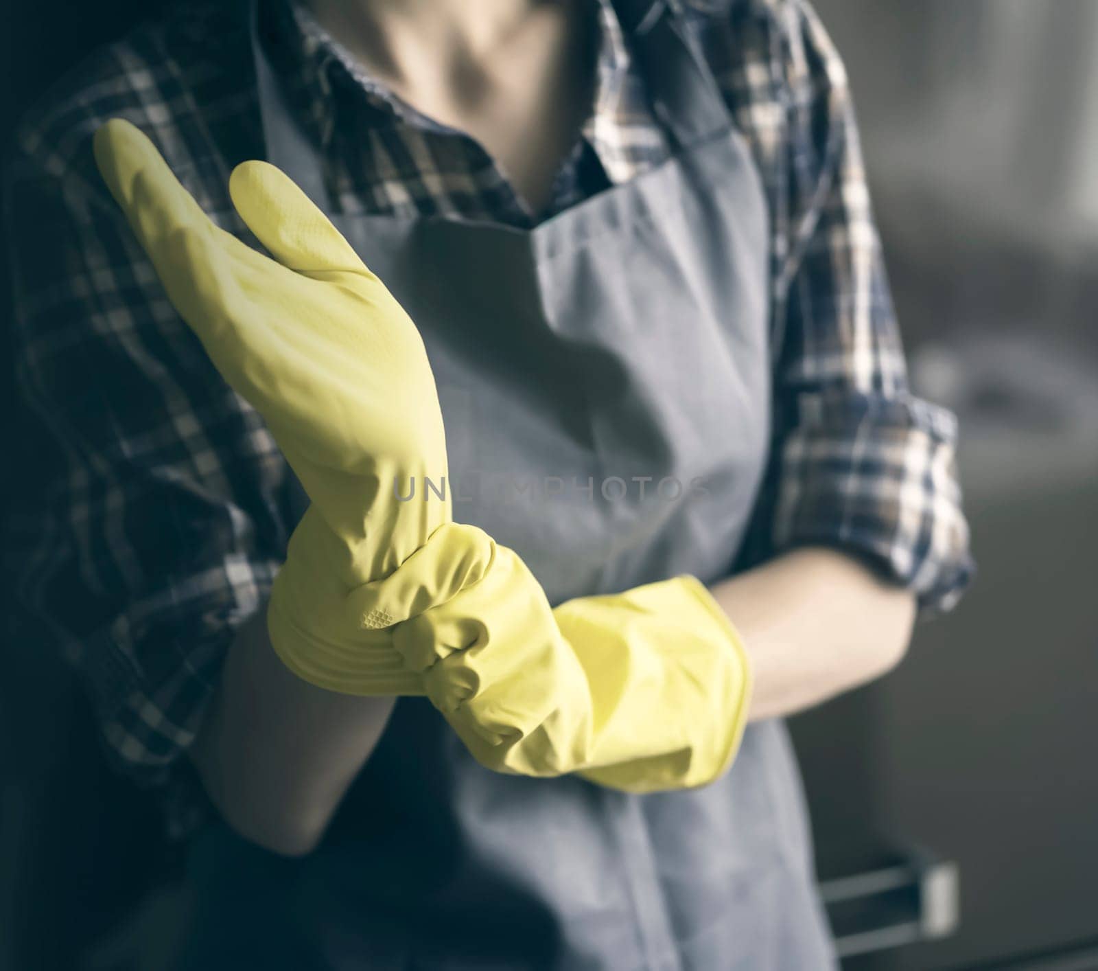 A young girl in a plaid shirt and apron puts yellow rubber gloves on her hands to start cleaning her house and create comfort. Housekeeper with gloves doing disinfection.