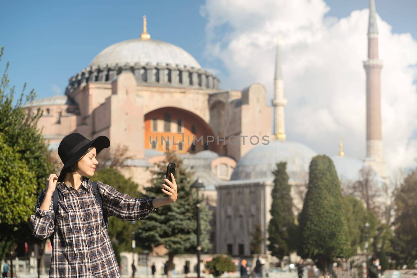 A young pretty girl in a stylish hat and a plaid shirt poses, makes a selfie on the phone near to the Hagia Sophia mosque. Tourism in Istanbul, Turkey.