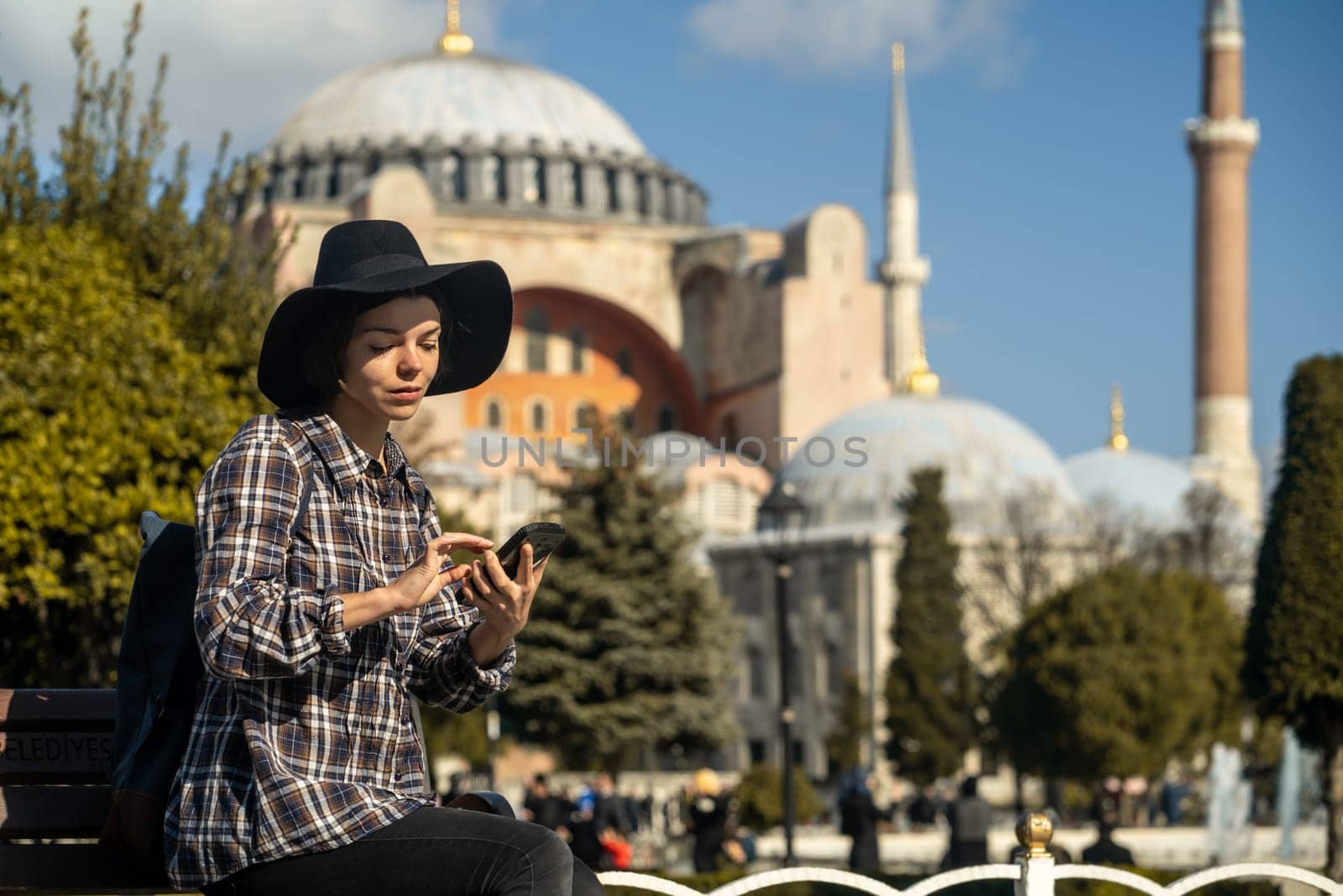 A young pretty girl in a stylish black hat and a plaid shirt sits on a bench and looks at the phone next to the Hagia Sophia mosque. Tourism in Istanbul, Turkey.