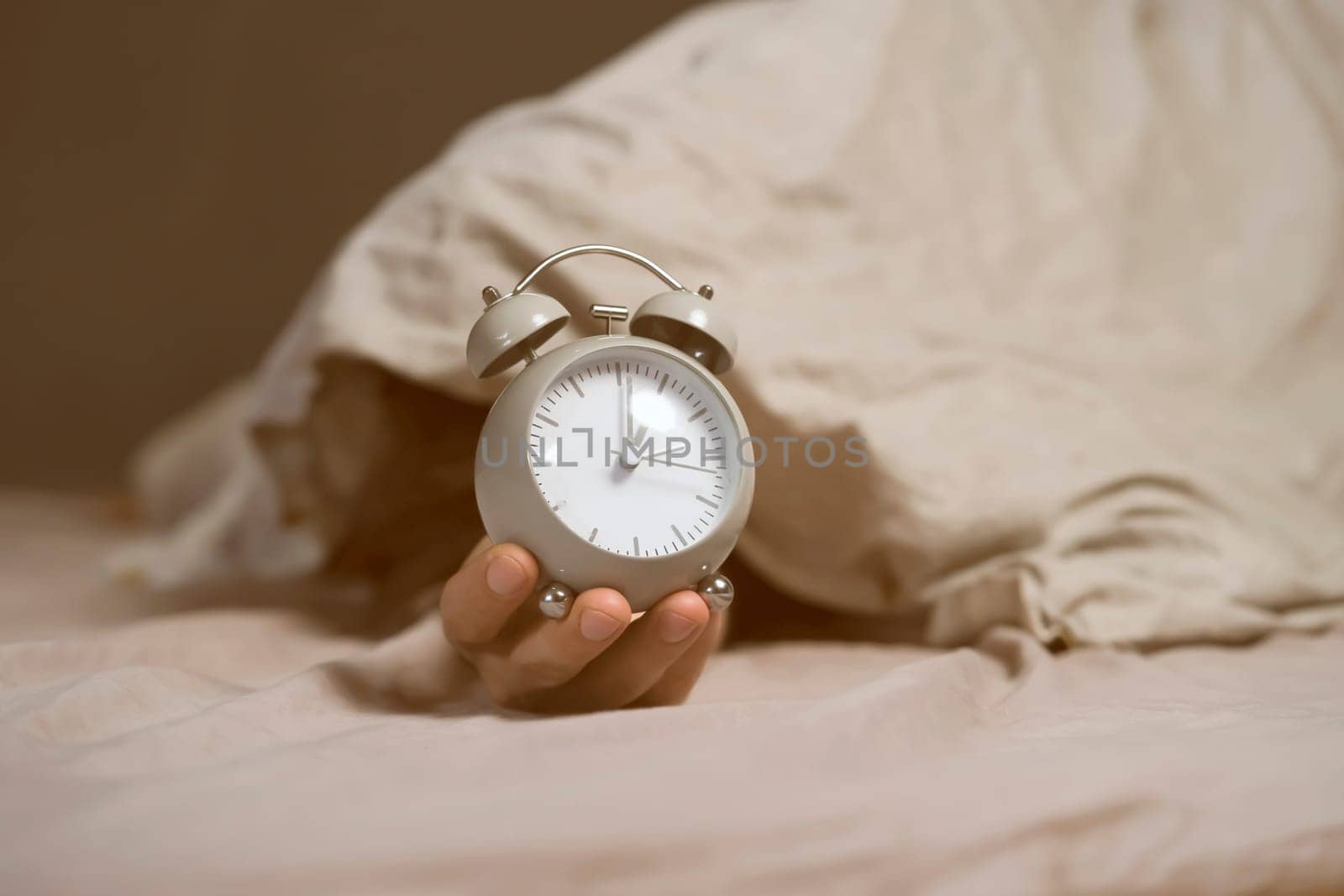 Hands of a young man from under the blankets hold a retro vintage alarm clock in gray. The person holds a clock, need to wake up.