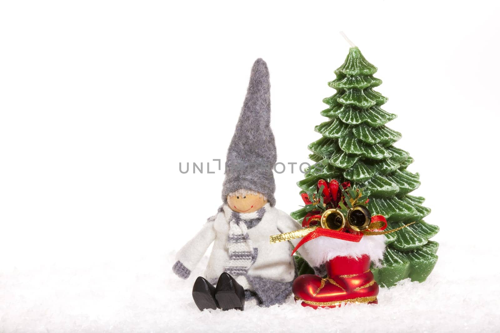 Little doll sitting next to red Santa boots and a candle Christmas tree in the snow against white background