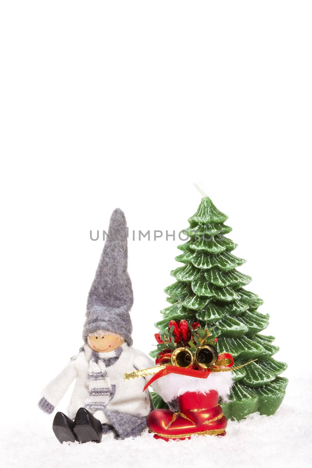 Little doll sitting next to red Santa boots and a candle Christmas tree in the snow against white background
