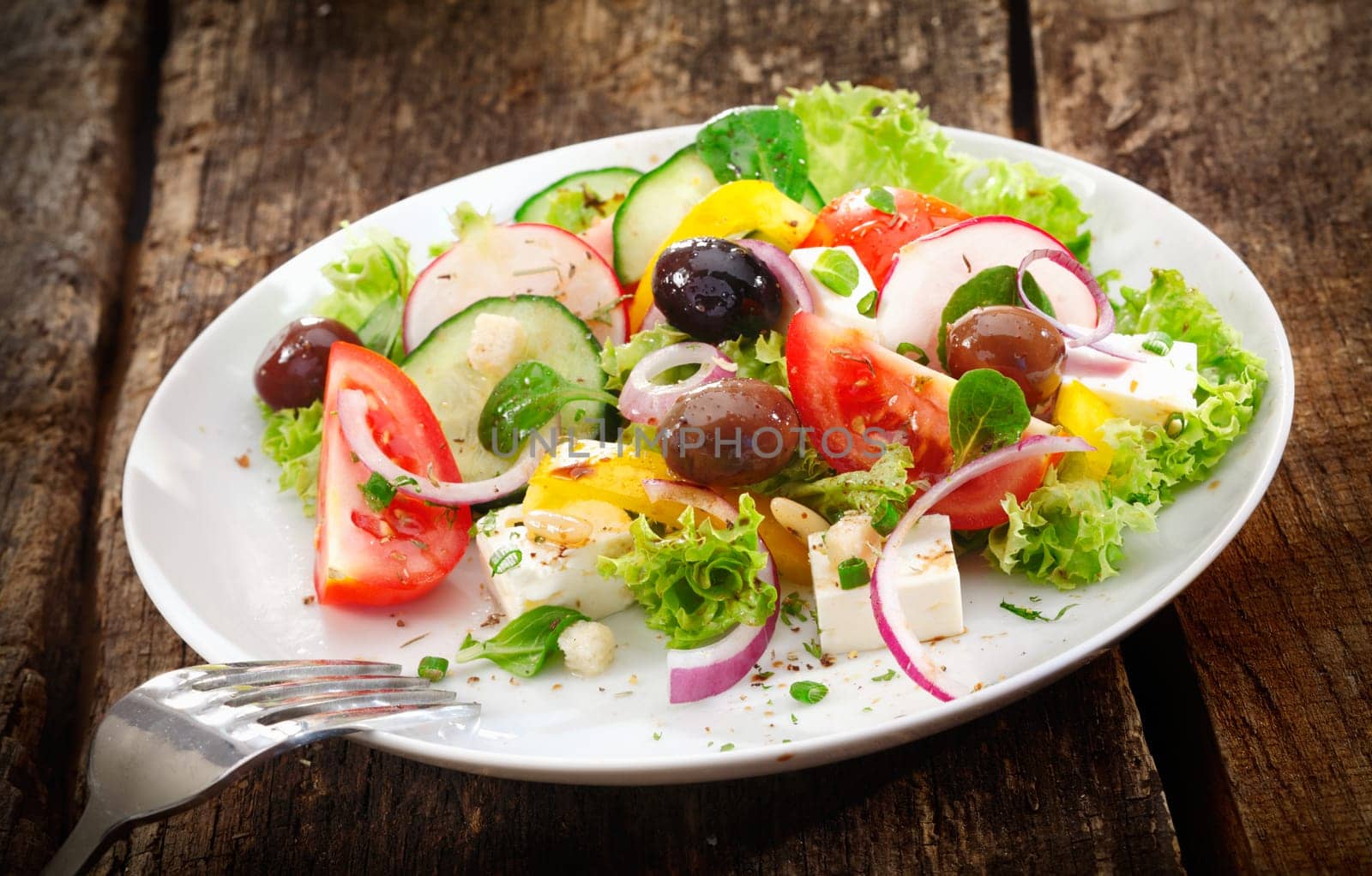 Serving of fresh healthy mixed salad with leafy greens, radish, tomato, olives and cheese served on a rustic wooden table