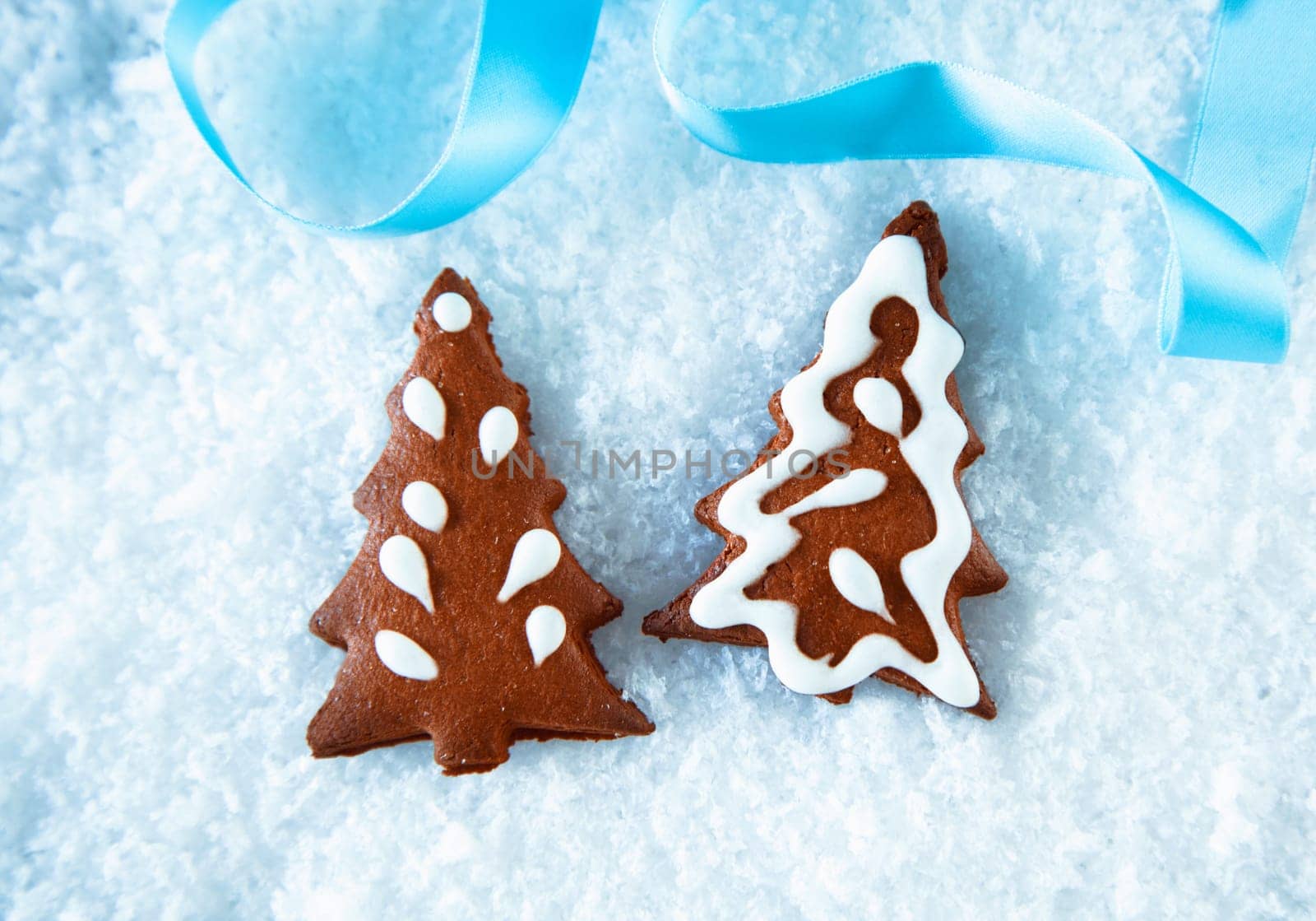 Crisp brown gingerbread Christmas trees with decorative icing lying on snow with a festive blue ribbon