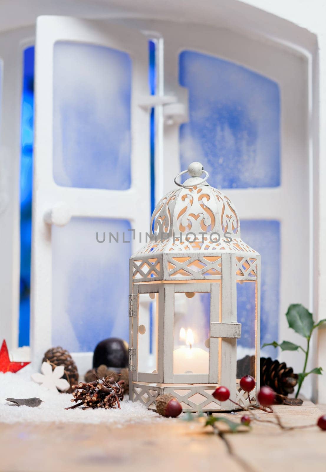 Pretty white Christmas lantern with a burning candle on a windowsill surrounded by decorative cones and berries