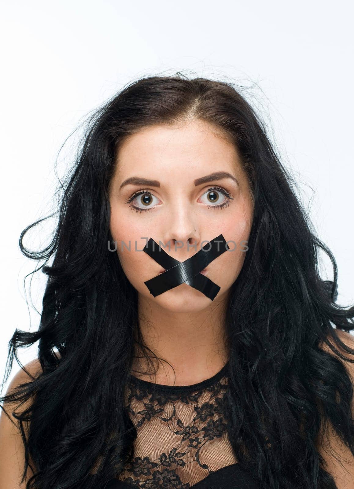 Head and shoulders shot of a young black-haired woman against a white background and mouth taped crosswise with black tape
