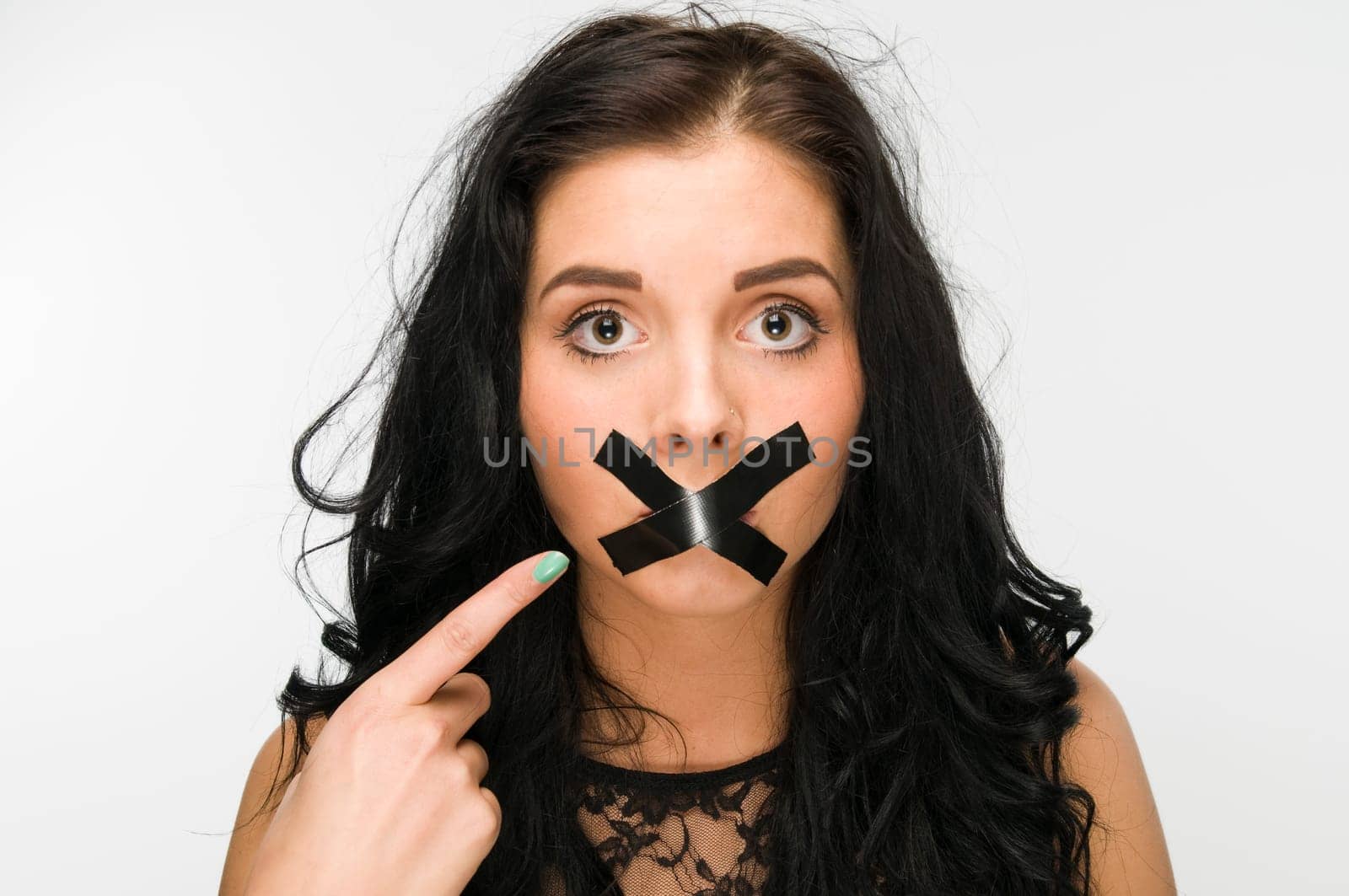 Head and shoulders shot of a young black-haired woman against a white background,mouth taped crosswise with black tape and pointing at the mouth with her index fingers