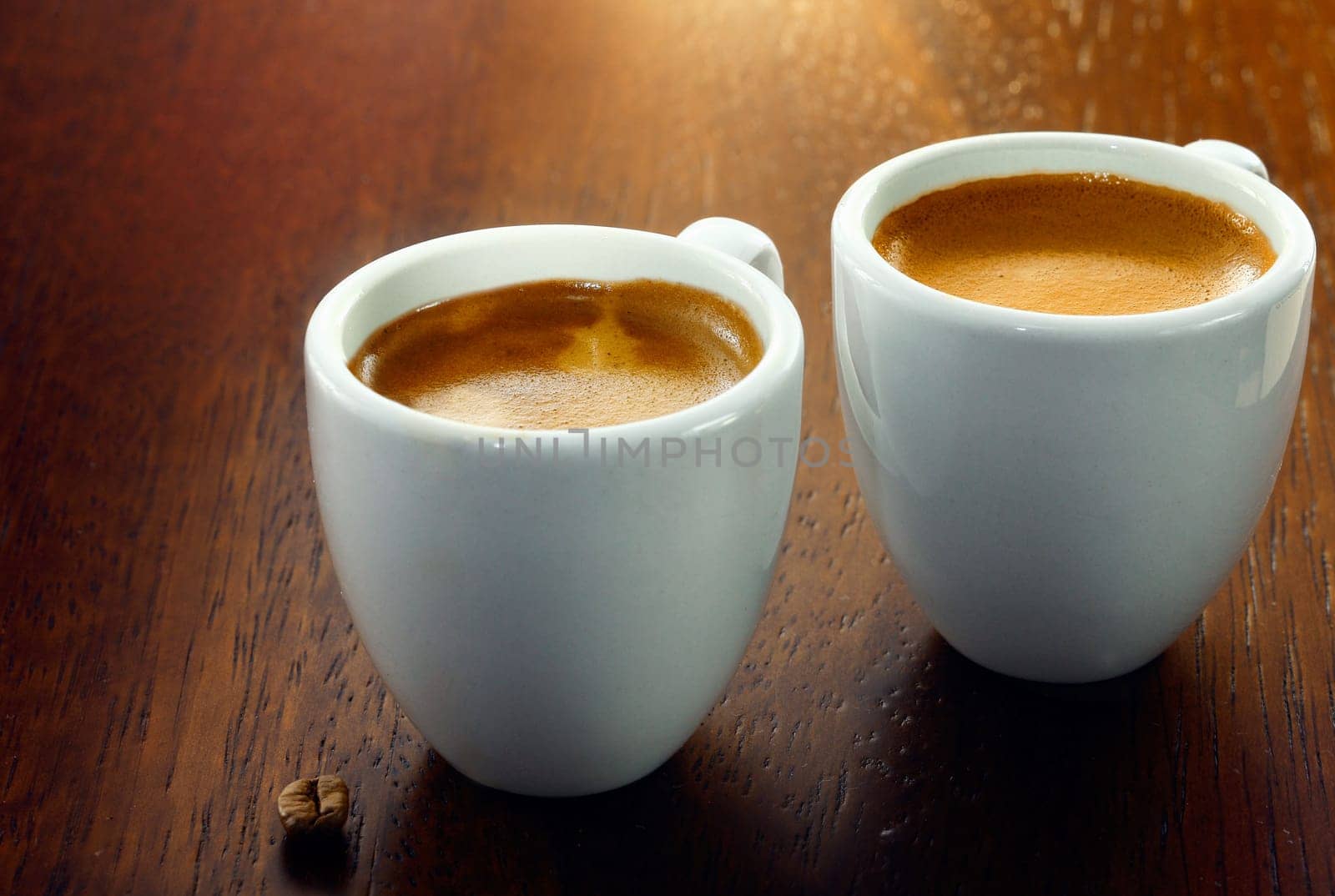 Two espresso coffees in small white cups,with a single coffee bean resting on the wood background