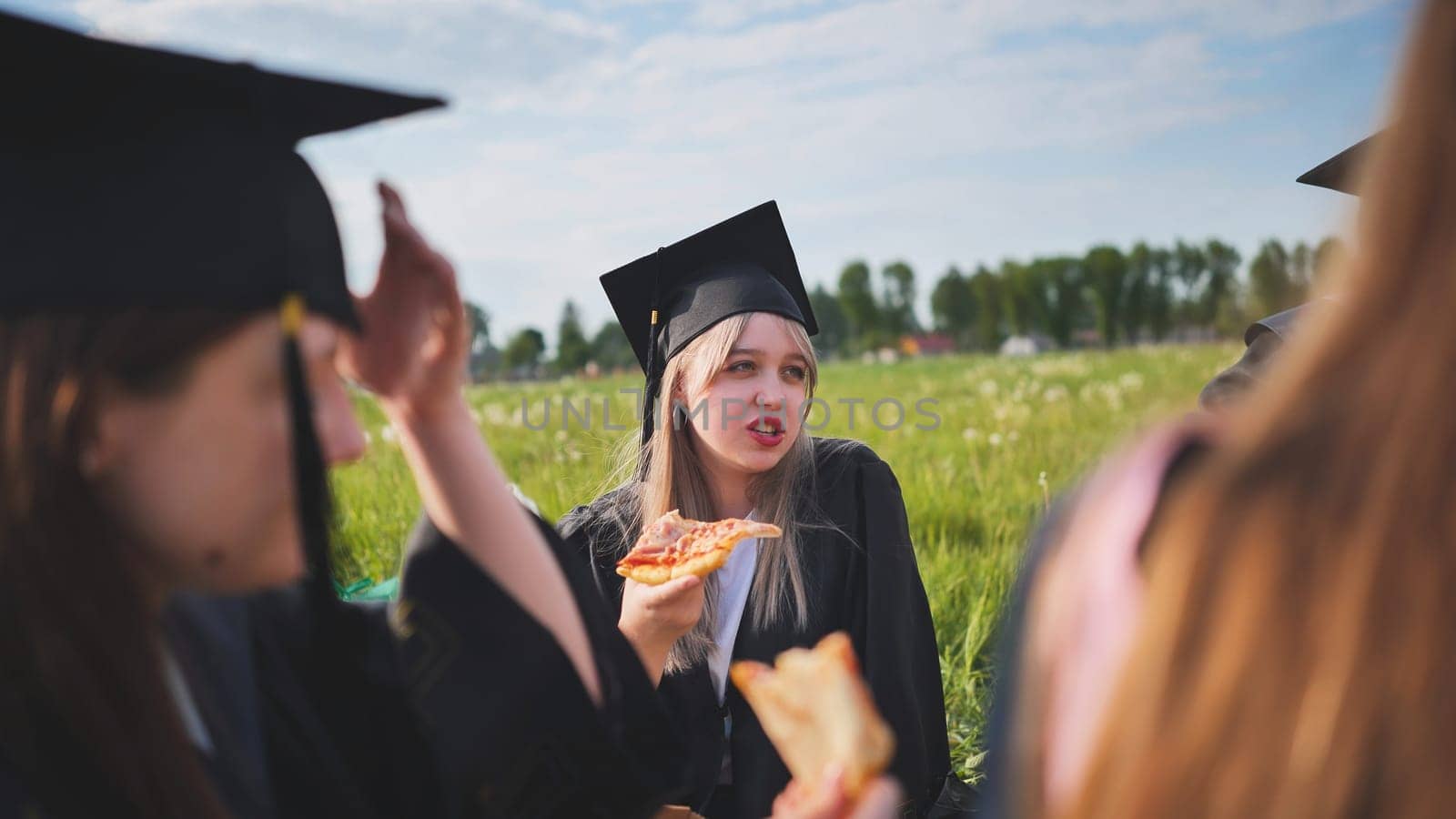 Graduates in black suits eating pizza in a city meadow