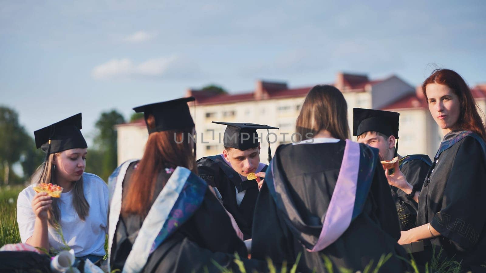 Graduates in black suits eating pizza in a city meadow. by DovidPro