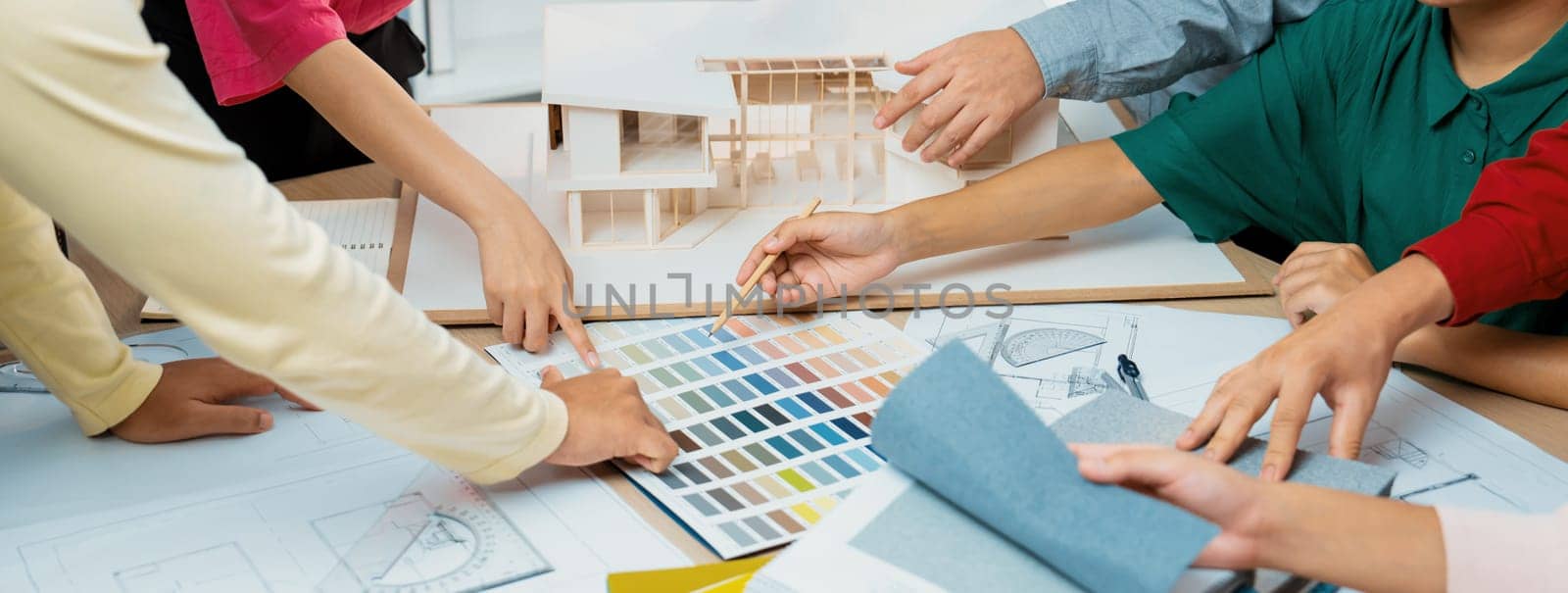 Skilled architect team discuss about house construction while interior designer point an appropriate color on color palette with house model and blueprint scatter around. Top view. Variegated.