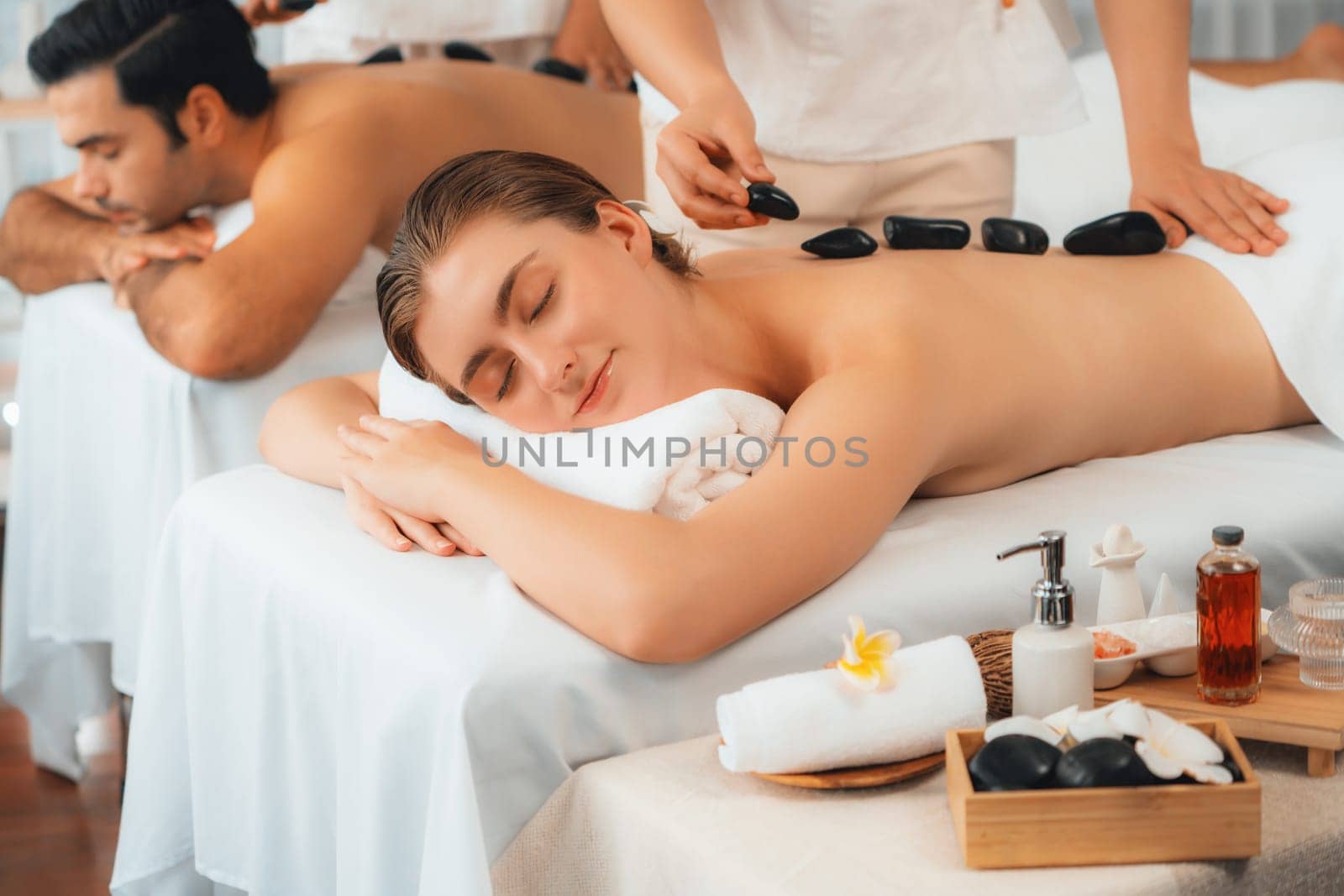 Hot stone massage at spa salon in luxury resort with day light serenity ambient, blissful couple customer enjoying spa basalt stone massage glide over body with soothing warmth. Quiescent