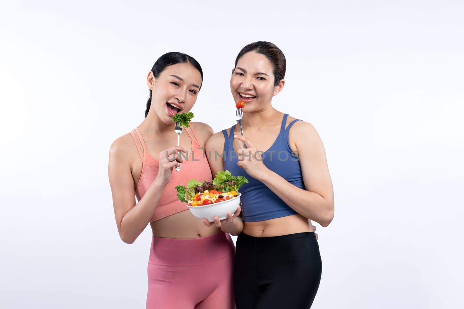 Two young sporty Asian women in sportswear holding salad bowl fill with fruit and vegetable. Natural youthful and fit body lifestyle people with balance nutrition on isolated background. Vigorous