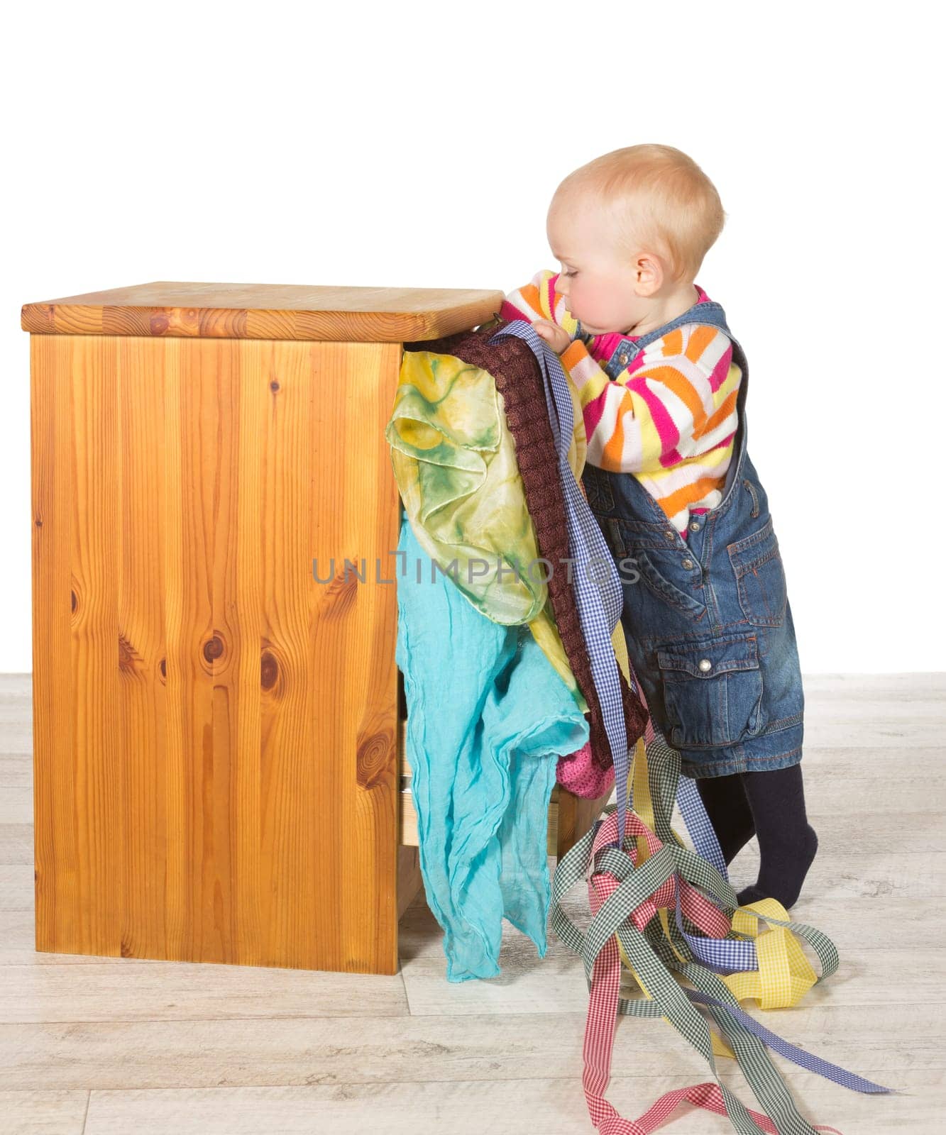 Determined toddler balancing on tiptoe stretching inside the top drawer of a small chest of drwawers to satisfy its curiosity as fabrics overflow onto the floor below