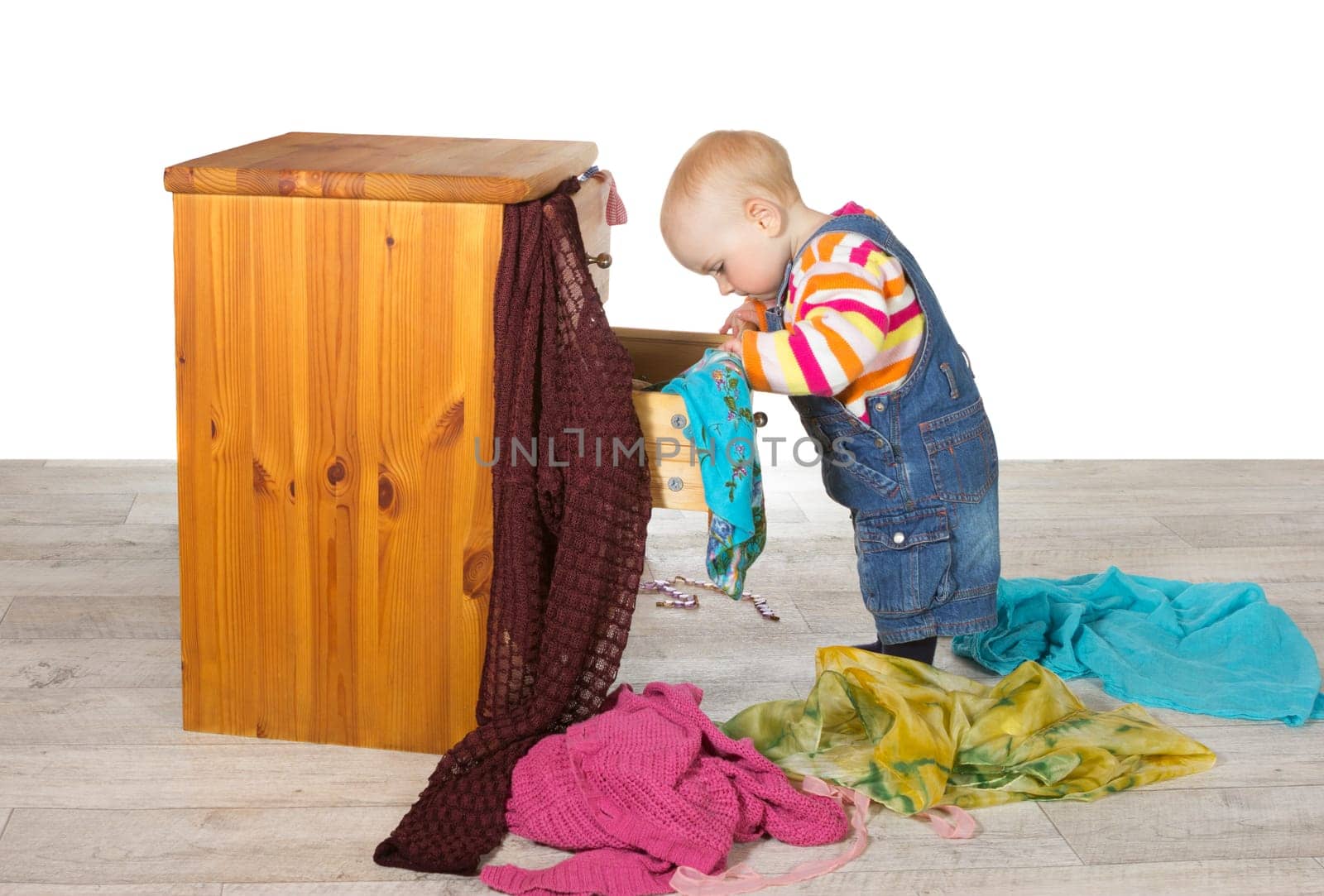 Happy baby standing searching in a drawer of a small wooden chest of drawers unpacking colourful clothes and textiles onto the floor in a random pile