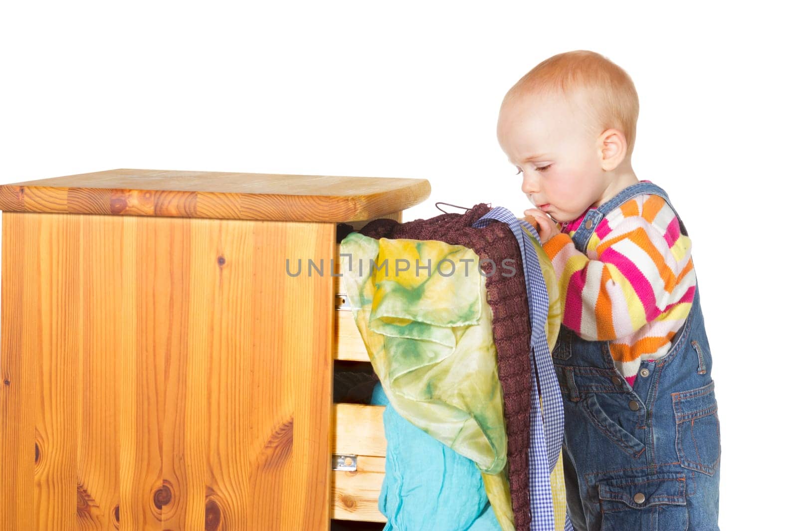 Curious little baby standing peering into a drawer with colourful textiles spilling out onto the floor on a white background