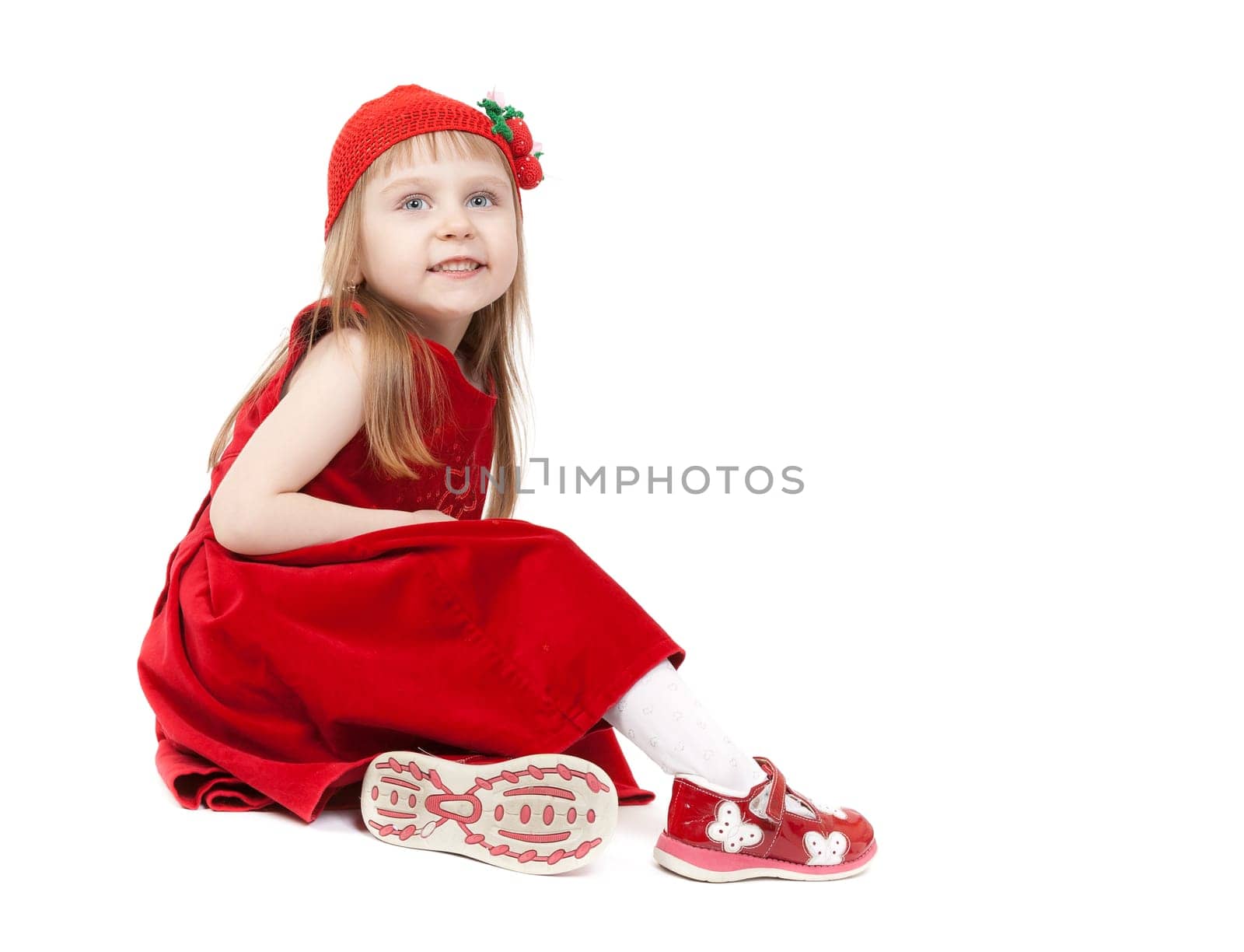 A little girl aged four years in a red dress and hat with strawberries sitting on the floor in the studio. Isolate on white