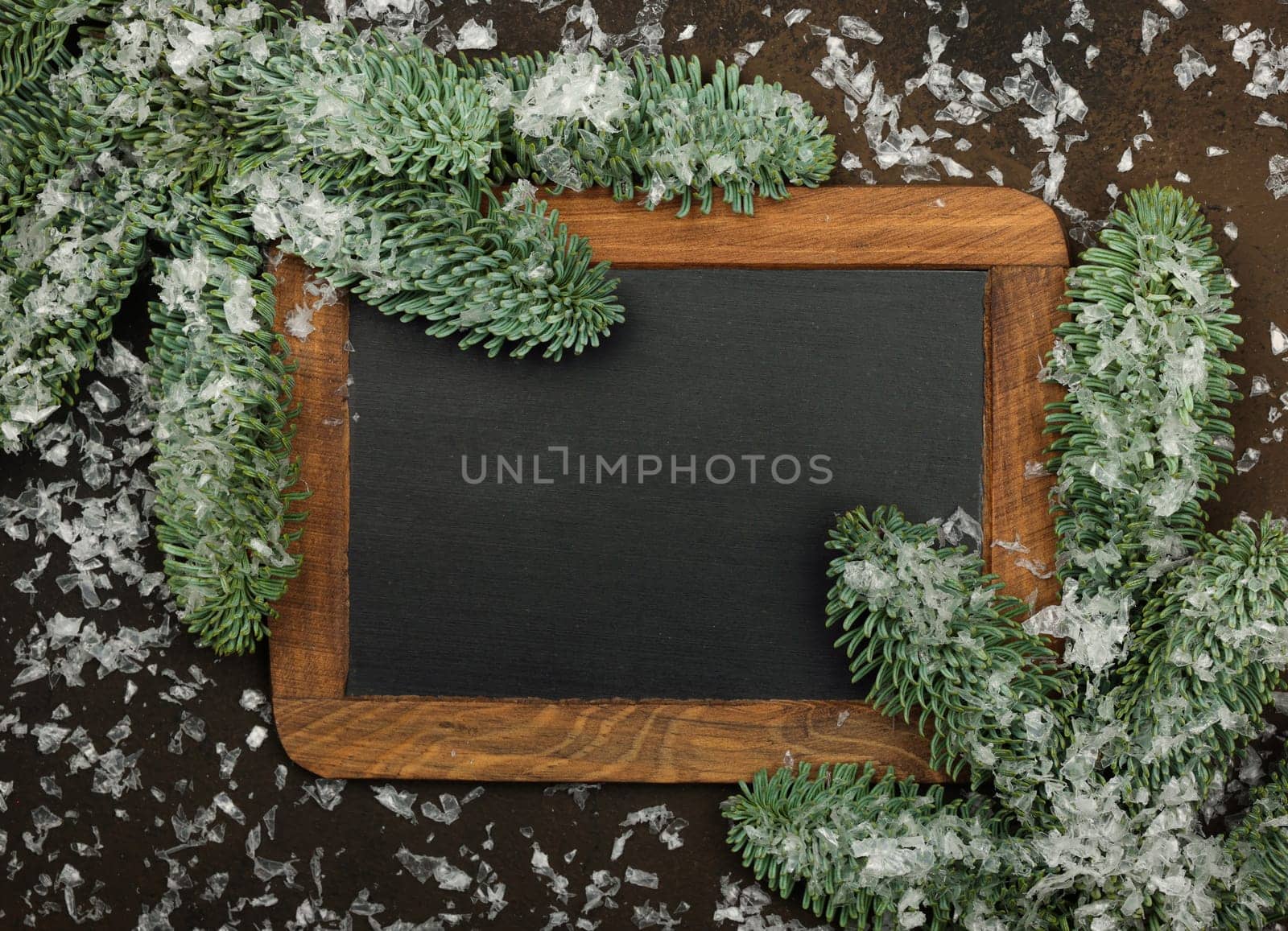 Close up vintage black chalkboard sign with wooden frame and Christmas decoration of fresh spruce branches and snow over dark brown background with copy space