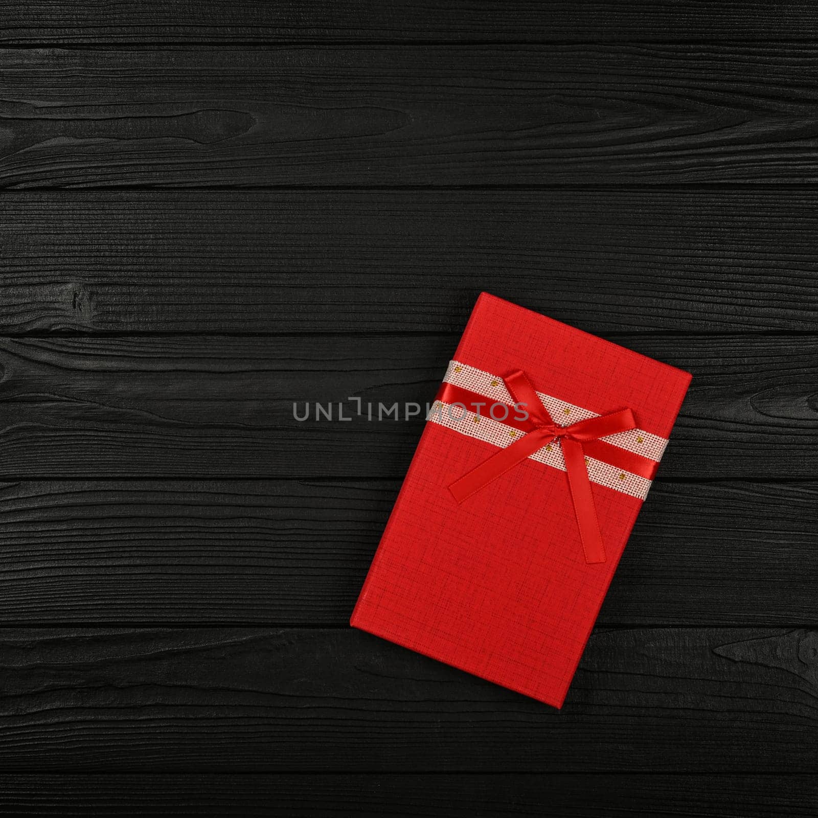 Valentine template of one closed vivid red gift box with ribbon bow over black wooden table background, close up flat lay, elevated top view, directly above