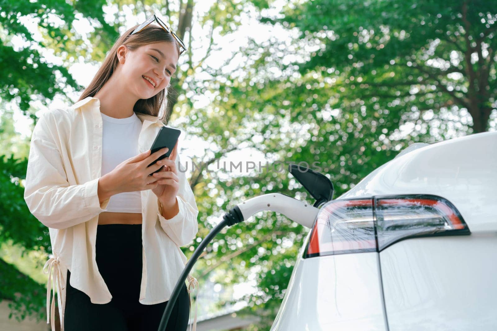 Young woman use smartphone to pay for electricity at public EV car charging station green city park. Modern environmental and sustainable urban lifestyle with EV vehicle. Expedient