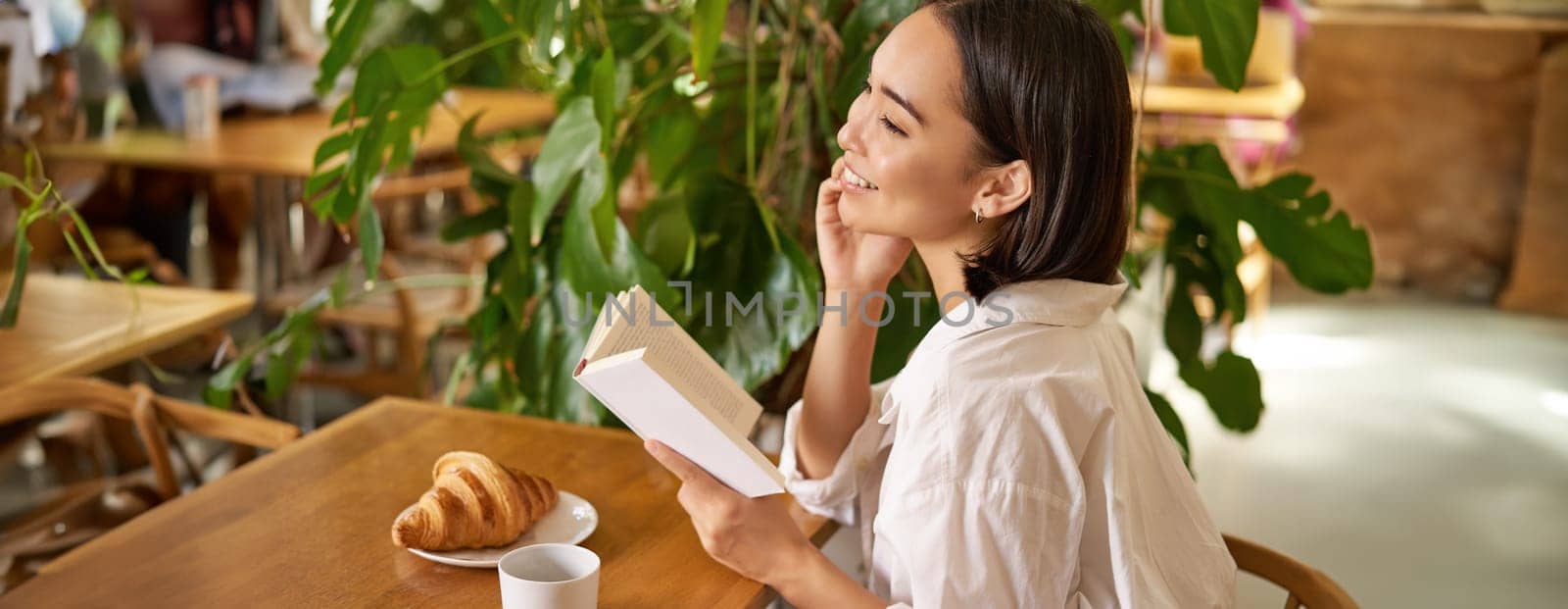 Beautiful young asian woman with a book in hands, sitting in cafe, drinking coffee and eating croissant, smiling, looking mysterious by Benzoix