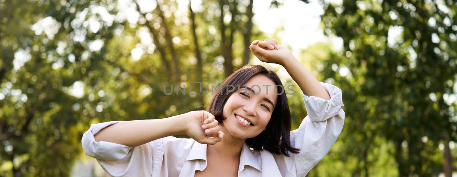 People lifestyle. Portrait of young brunette woman dancing, smiling and laughing, walking in park with hands lift up high, enjoying summer day outside by Benzoix