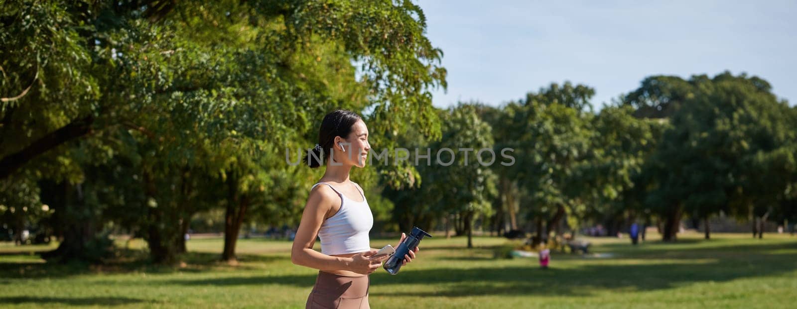 Active asian girl, in fitness clothing, workout in park, walking in sportswear with smartphone and water bottle by Benzoix