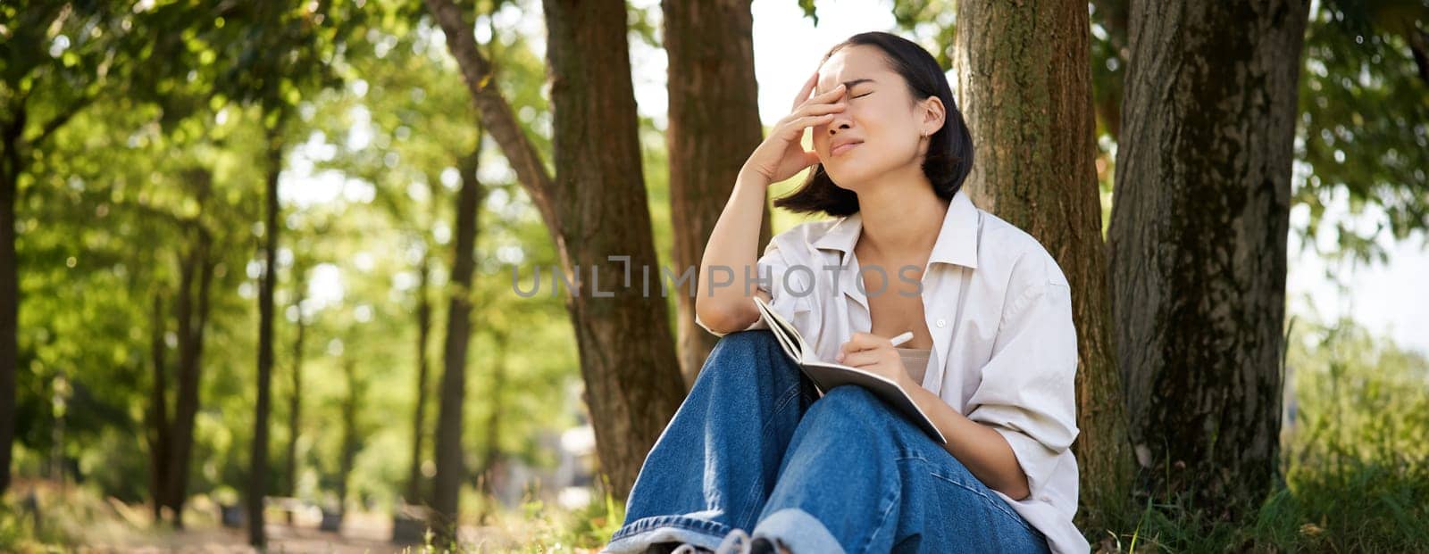 Portrait of sad asian girl writing in her diary and feeling uneasy, sitting in park alone under tree, expressing her distress in notebook.