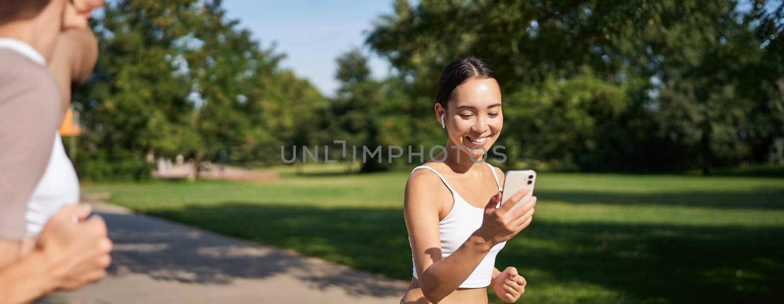 Happy asian fitness girl running and checking her stats, daily goals on smartphone app. Young woman jogging and looking at mobile screen.