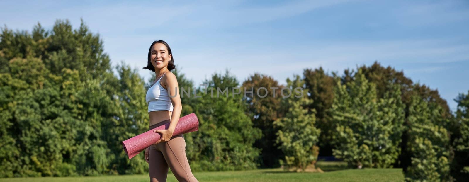 Vertical shot of stylish fitness woman with rubber mat, posing on lawn in park, doing workout on fresh air by Benzoix