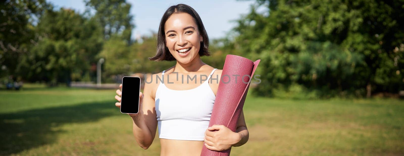 Smiling asian fitness girl with rubber yoga mat, shows her smartphone screen, recommends workout application, stands on lawn in park.