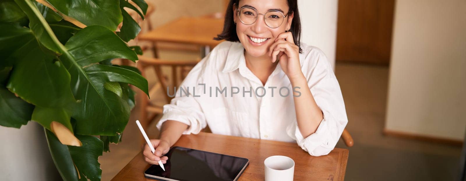 Young asian creative girl, sitting with digital tablet, drawing with graphic pen and smiling, doodling, drinking coffee in cafe.