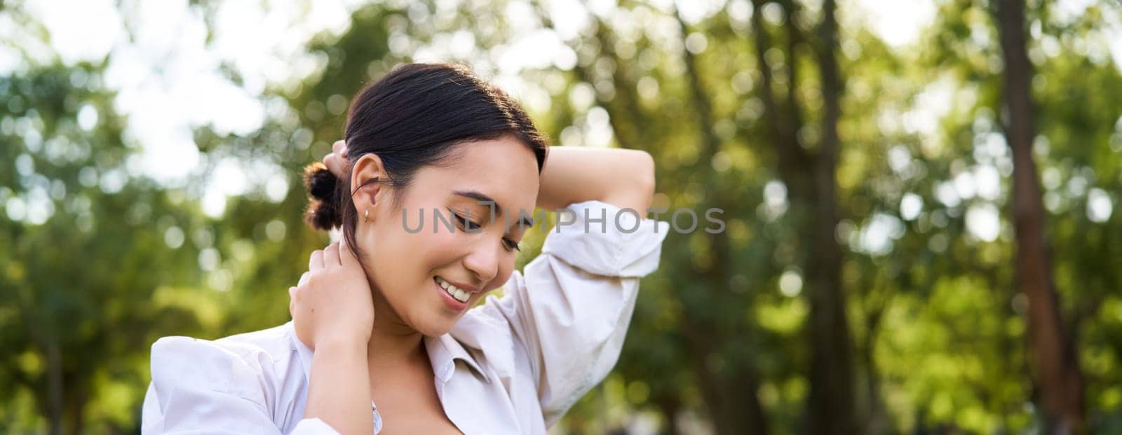 Beautiful young woman tying her hair while walking in park, smiling romantic, enjoying warm day.