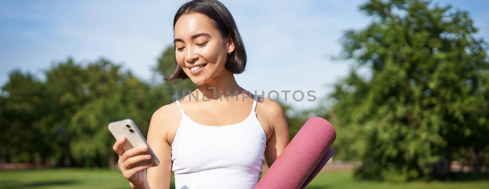 Portrait of smiling asian woman with yoga mat, looking at her smartphone and reading on application, standing in park wearing sport uniform by Benzoix