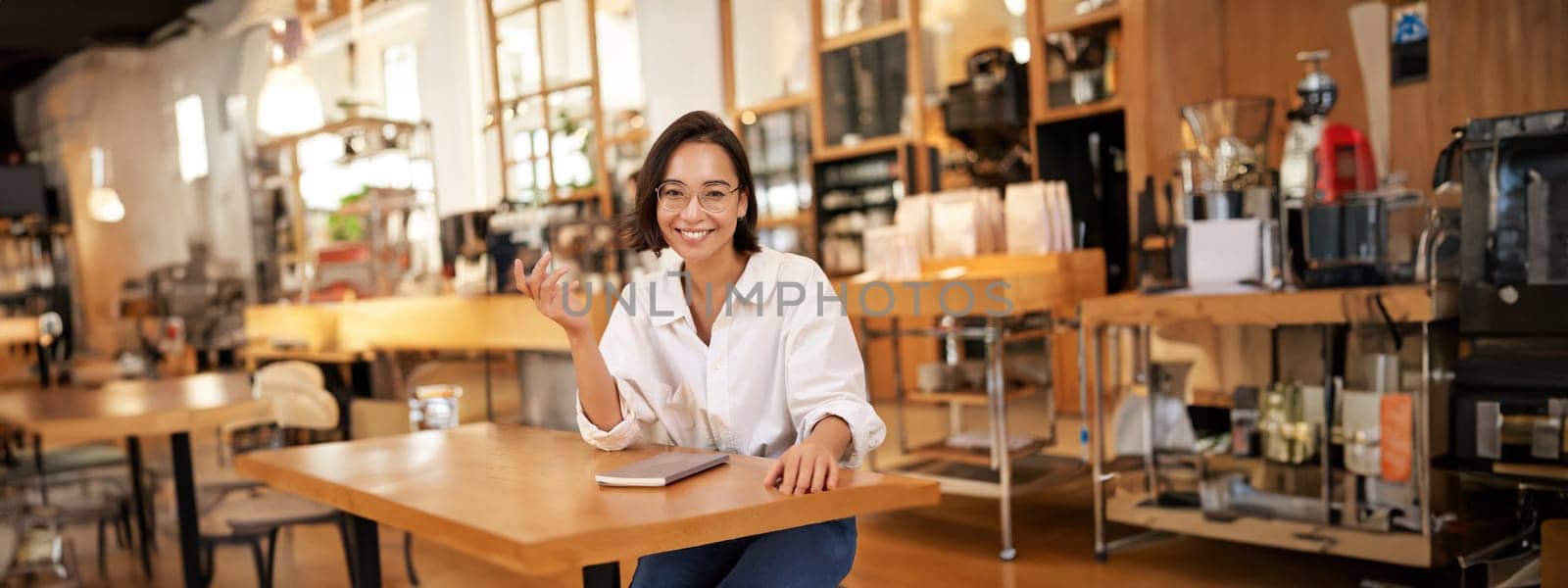 Young stylish asian woman, business owner in glasses, sitting in cafe with notebook, smiling at camera. People and lifestyle concept