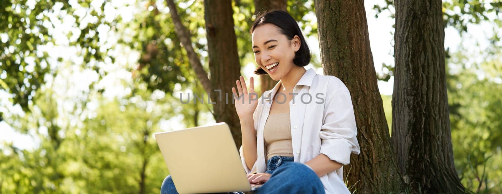 Happy young asian girl sits in park near tree, looking at laptop, working remotely from outdoors, talking to someone, video chat.