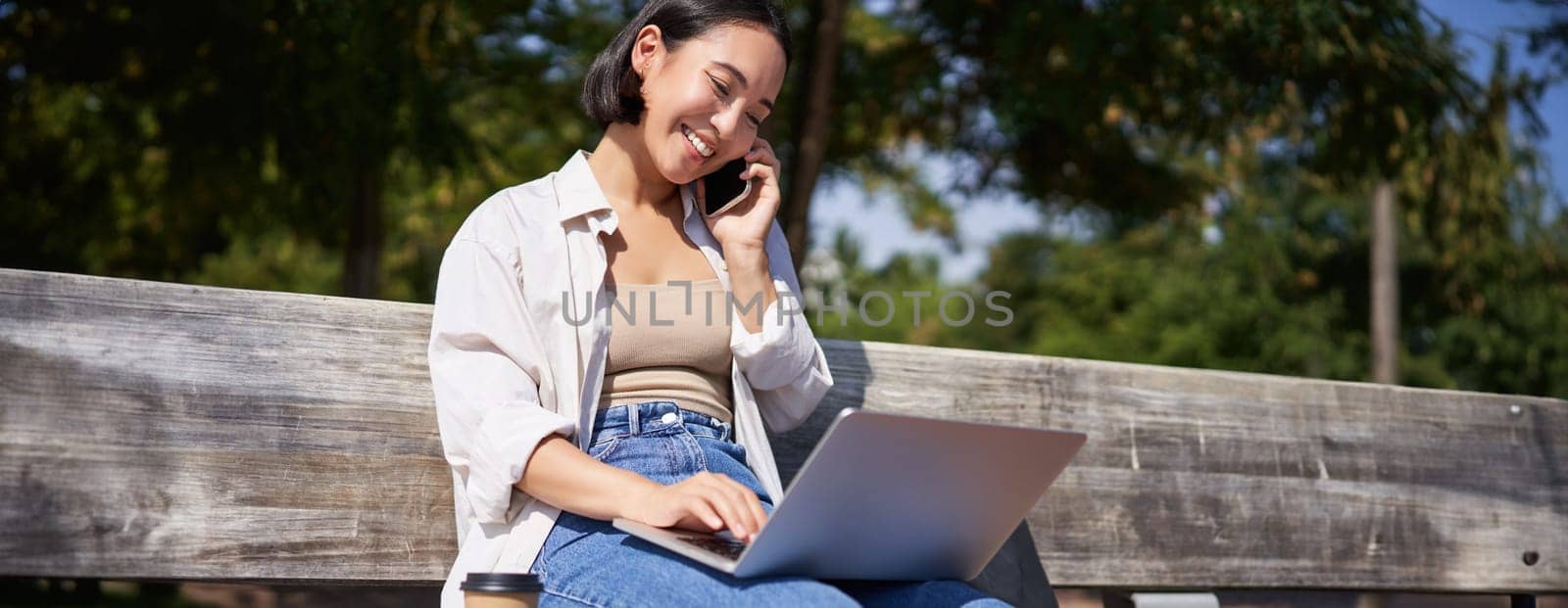 Young asian girl talking on mobile phone, using laptop, working online from park, sitting on bench on sunny day with computer by Benzoix