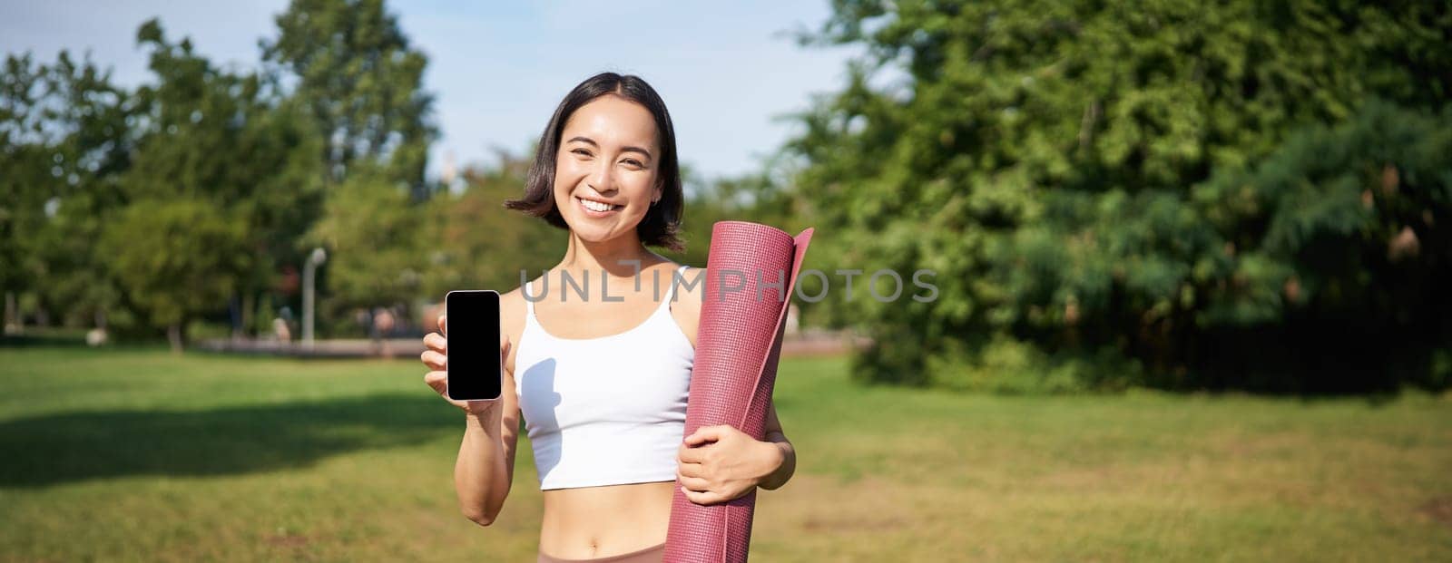 Excited fitness girl recommends application for sport and workout, shows phone screen, standing with rubber yoga mat in park after training session by Benzoix