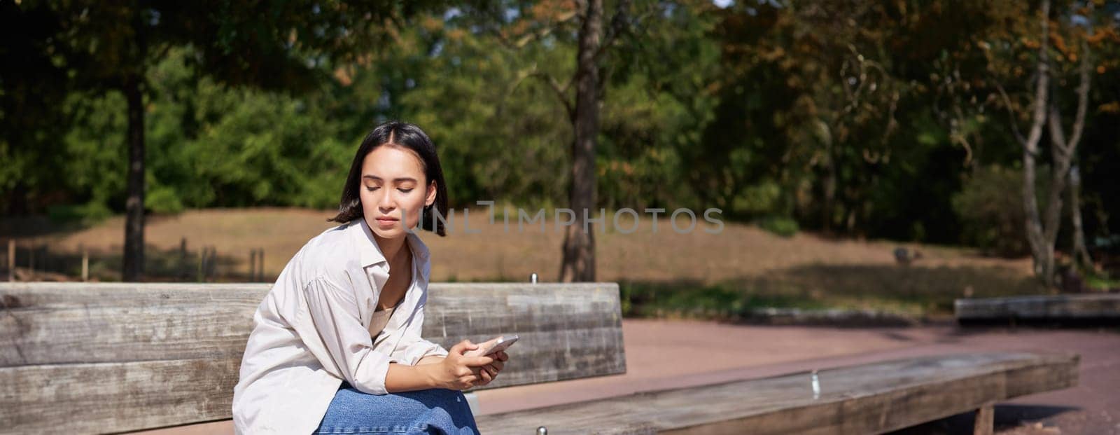 Portrait of asian girl sitting with smartphone feeling sad, looking gloomy and frustrated, waiting for a call outdoors in park by Benzoix