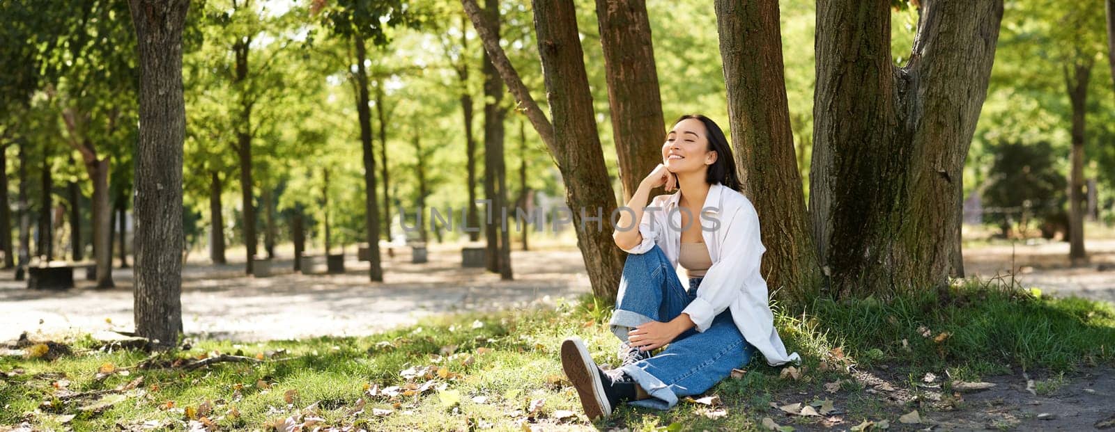Relaxed young woman, resting near tree, sitting in park on lawn under shade, smiling and looking happy, walking outdoors on fresh air by Benzoix