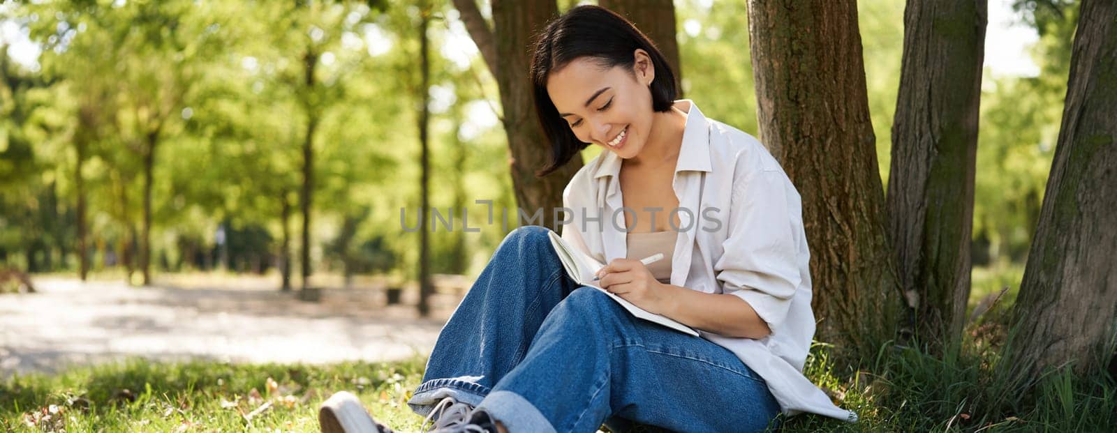 Portrait of happy asian girl sitting in park and writing in her diary. Young woman doing homework on fresh air, sitting near tree and smiling by Benzoix