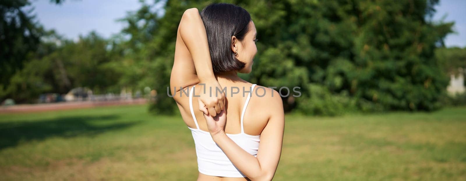 Rear view of young sporty woman stretching her arms behind back, warm-up, prepare for workout jogging, sport event in park.